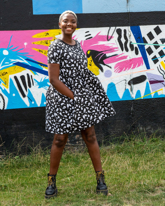 Chish models a stretch cotton smock dress featuring an all-over white ghost print. This alternative casual dress has short sleeves, side pockets, and a relaxed fit, ideal for everyday gothic and quirky style.