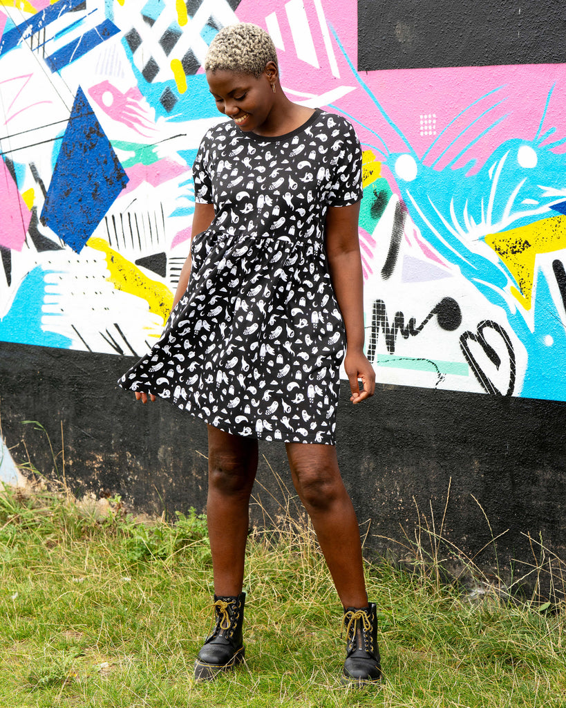 Chish wearing the Rebel Romance Ghosties Smock Dress, a black stretch cotton dress with white ghost prints. She poses outdoors, holding the skirt slightly to the side to show the relaxed smock fit and short sleeves.
