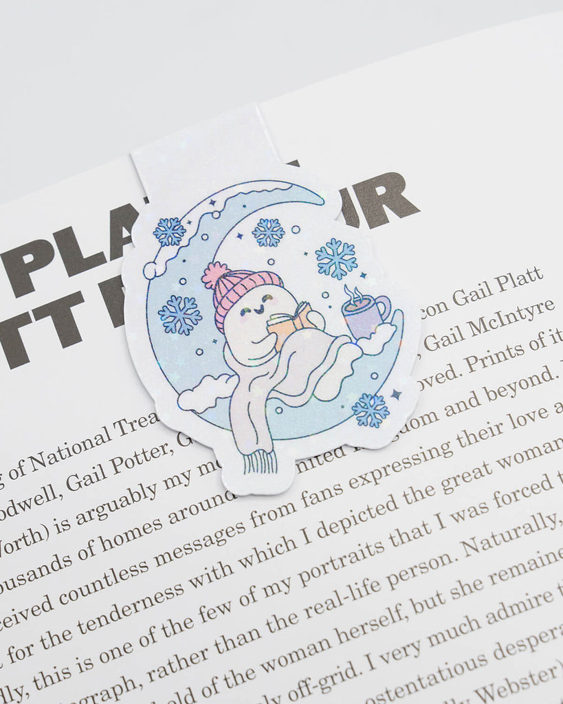 Magnetic bookmark clipped to a book page featuring a cute ghost wrapped in a blanket, reading a book while sitting on a crescent moon. Surrounded by snowflakes and a steaming mug of cocoa, this pastel design is perfect for winter reading vibes. A cosy and whimsical gift for book lovers, exclusively from Rebel Romance.