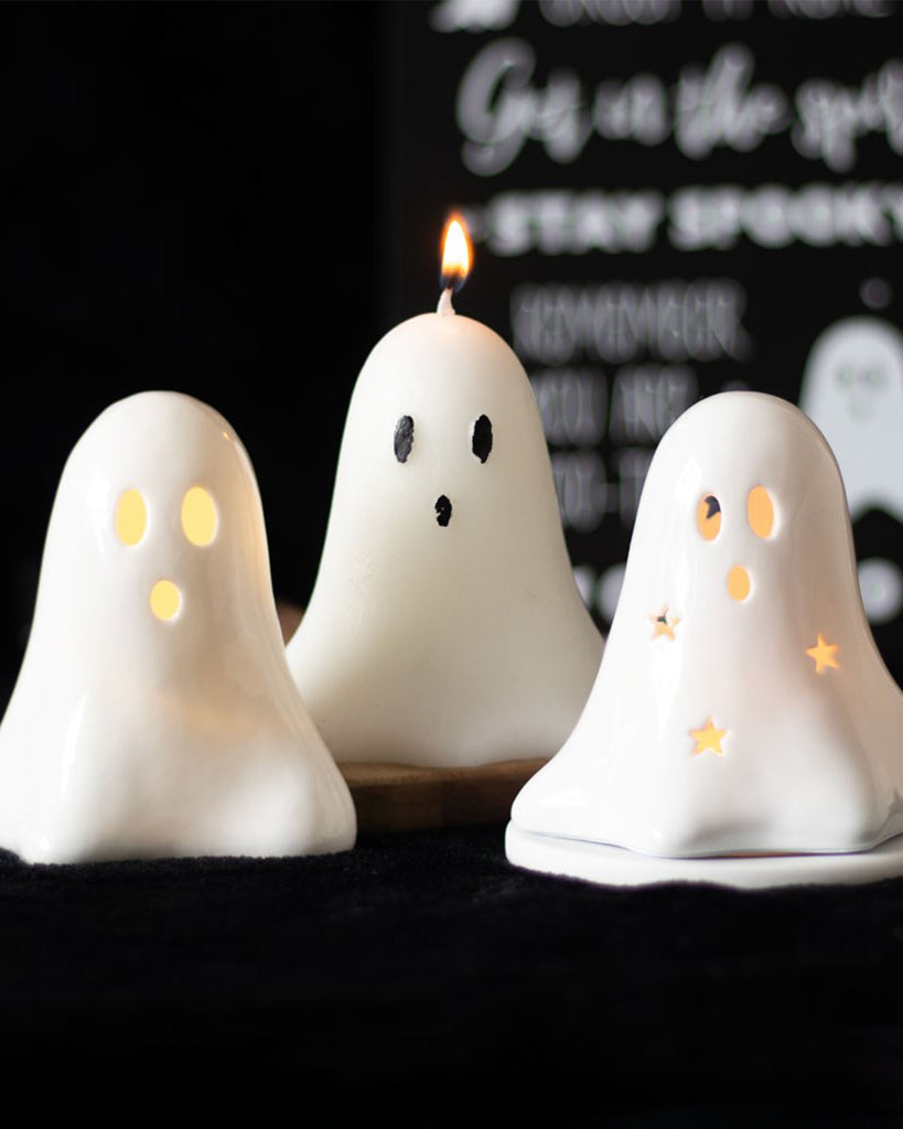 Three ghost-shaped Halloween decorations on a black surface with glowing lights and a spooky candle. The central ghost is a lit unscented candle, while the others are decorative ghost lights with star and circle cutouts. Perfect for gothic or alternative home decor from Rebel Romance.