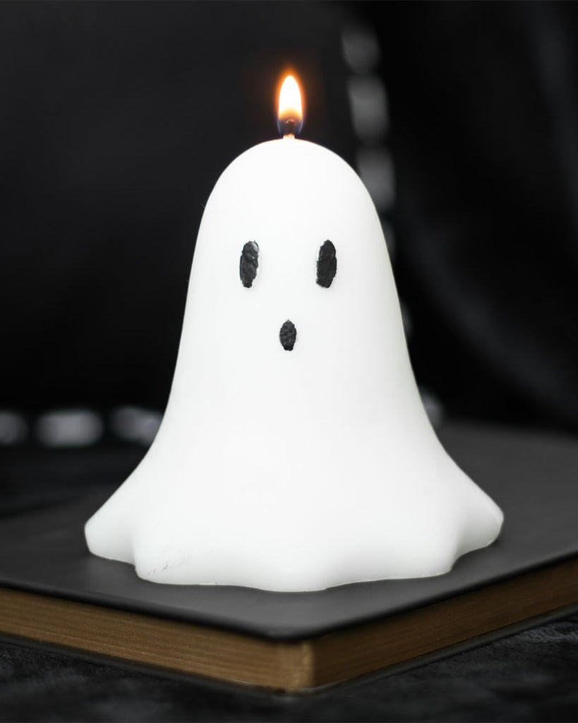 Rebel Romance Unscented Ghost Candle lit and glowing with a flame. The candle sits on a dark book, making it a moody and stylish Halloween home decor piece for alternative and spooky interiors.