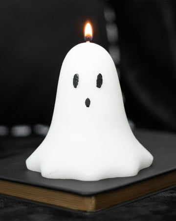 Rebel Romance Unscented Ghost Candle lit and glowing with a flame. The candle sits on a dark book, making it a moody and stylish Halloween home decor piece for alternative and spooky interiors.