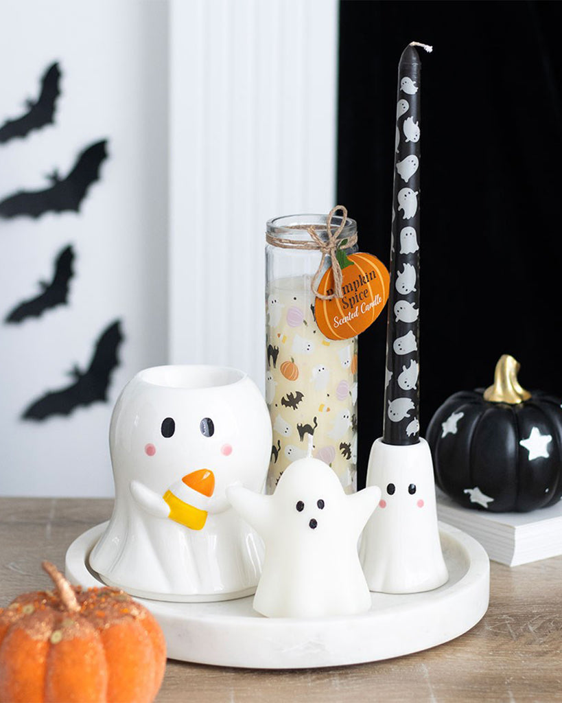 A Halloween-themed tabletop display featuring the Rebel Romance Sweet Ghost Candle Holder with a black taper candle decorated in white ghost illustrations. Surrounded by a mini pumpkin, ghost-shaped decor, a pumpkin spice candle, and a black ceramic pumpkin, set against a backdrop with black paper bats on the wall.