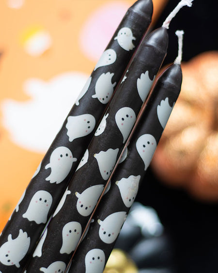 Three ghost-themed taper candles by Rebel Romance held together in a Halloween setting with pumpkins in the background. The cute white ghost pattern on black makes these a charming yet gothic addition to your seasonal décor or dinner table.