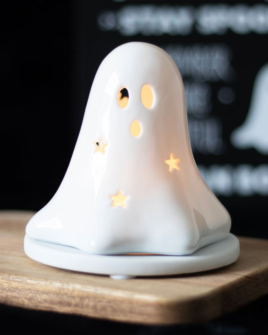A glowing ceramic ghost tealight holder placed on a wooden board in a dim setting, adding warmth and spooky charm to your space. Sold by Rebel Romance UK, this versatile incense or candle holder makes a bewitching home accessory.