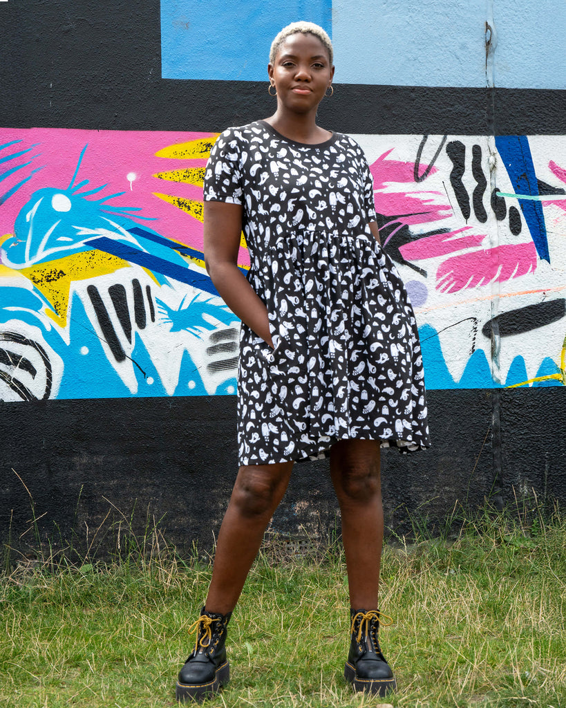 Chish wearing the Rebel Romance Ghosties Smock Dress, a black stretch cotton dress with white ghost prints. She poses outdoors, holding the skirt slightly to the side to show the relaxed smock fit and short sleeves.
