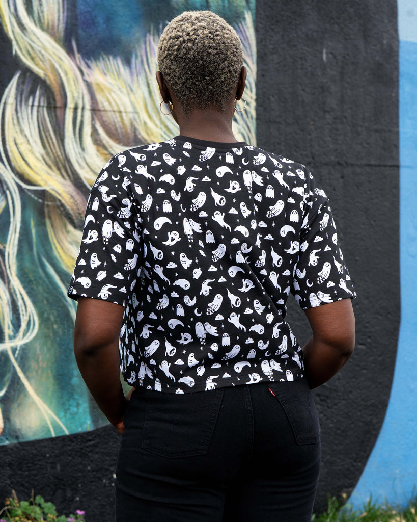 Back view of Chish wearing a black t-shirt with white ghosties illustrated by Yasmin Cousins against a colourful mural background