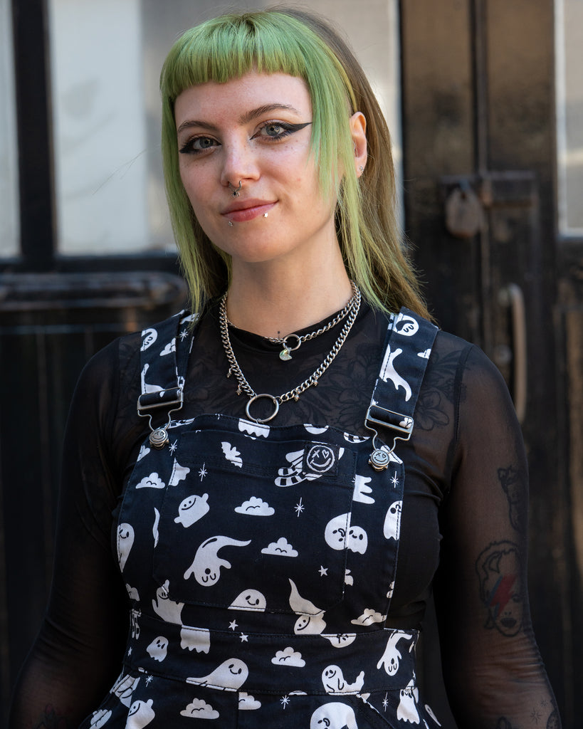 Close up of a femme person with green hair twirling smiling t patterned flared pinafore dress with a mesh long sleeve top  in front of a black door. Design by Yasmin cousins. 