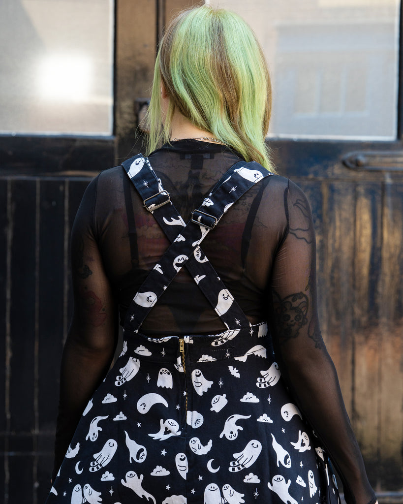 Close up of a femme person with green hair facing away wearing a black and white ghost patterned flared pinafore dress with a mesh long sleeve top in front of a black door. Design by Yasmin cousins. 