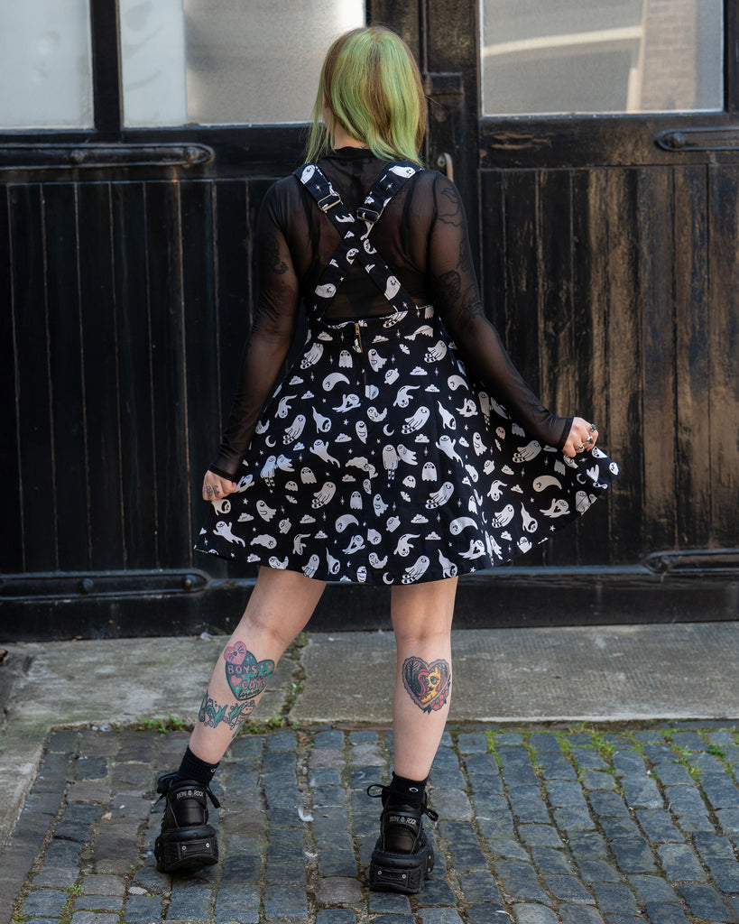 Femme person with green hair facing away wearing a black and white ghost patterned flared pinafore dress with a mesh long sleeve top and black shoes on a cobbled street. Design by Yasmin cousins. 