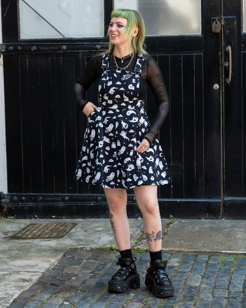 Femme person with green hair laughing wearing a black and white ghost patterned flared pinafore dress with a mesh long sleeve top and black shoes on a cobbled street. Design by Yasmin cousins. 