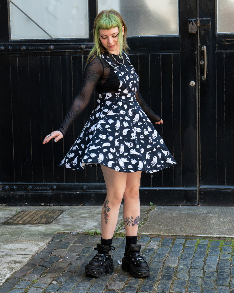 Femme person with green hair twirling wearing a black and white ghost patterned flared pinafore dress with a mesh long sleeve top and black shoes on a cobbled street. Design by Yasmin cousins. 