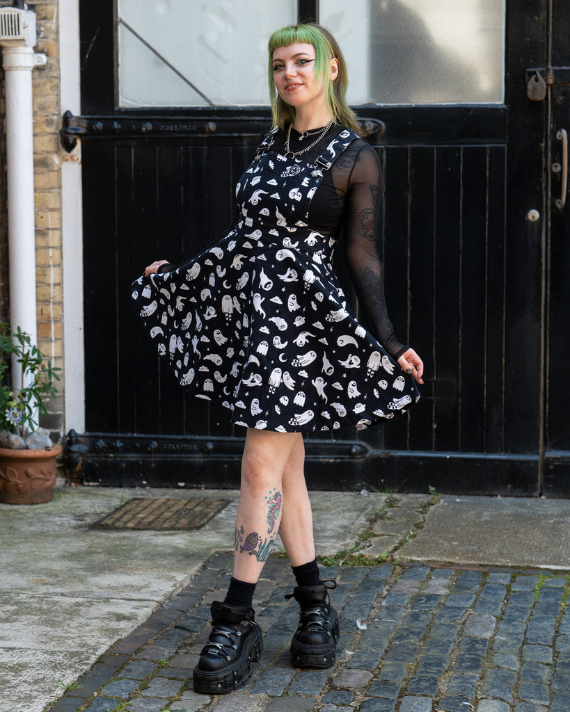 Femme person with green hair smiling wearing a black and white ghost patterned flared pinafore dress with a mesh long sleeve top and black shoes on a cobbled street. Design by Yasmin cousins.