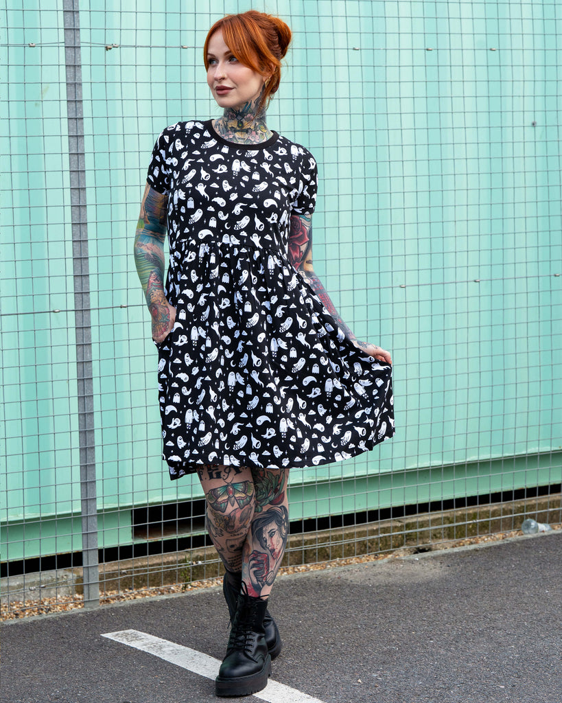 Erin wearing the Rebel Romance Ghosties Smock Dress, a black stretch cotton dress with white ghost prints. She poses outdoors, holding the skirt slightly to the side to show the relaxed smock fit and short sleeves.
