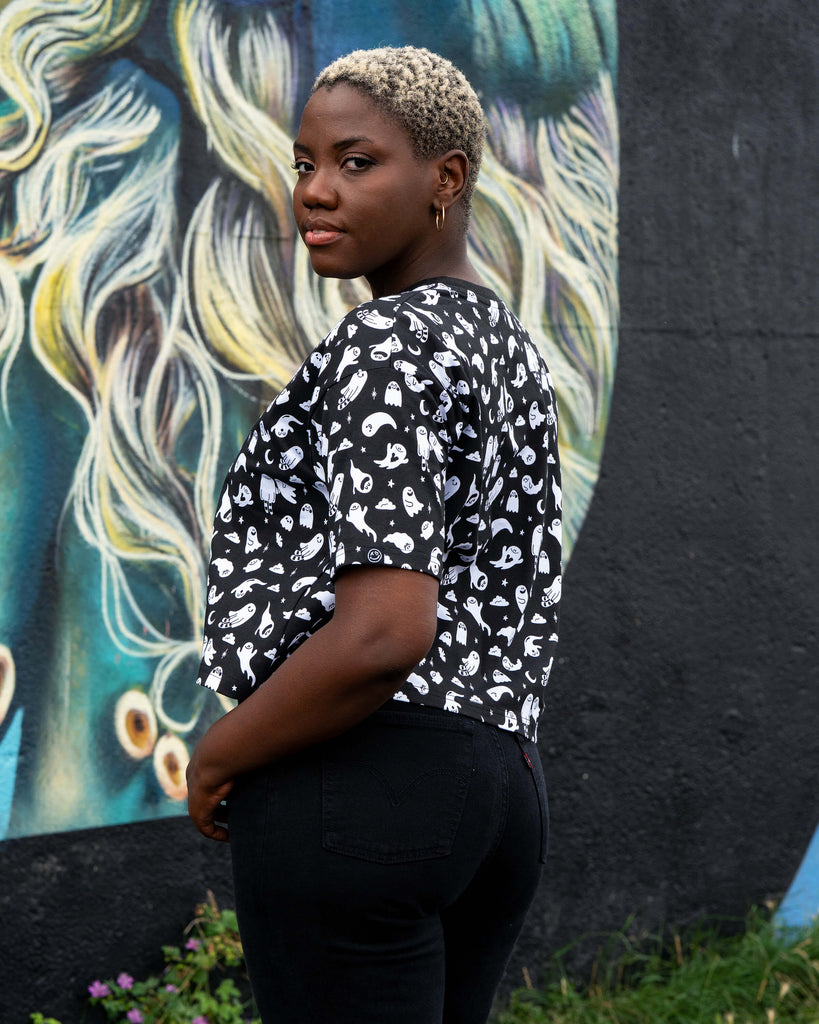 Chish wearing a black t-shirt with white ghosties illustrated by Yasmin Cousins against a colourful mural background. She is stood to the side looking over her shoulder.