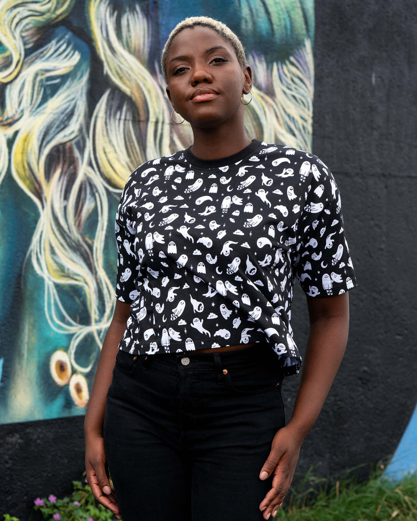 Chish wearing a black t-shirt with white ghosties illustrated by Yasmin Cousins against a colourful mural background