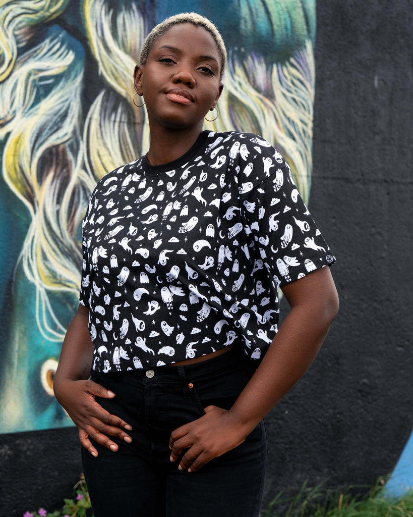 Front view of Chish wearing a black t-shirt with white ghosties illustrated by Yasmin Cousins against a colourful mural background