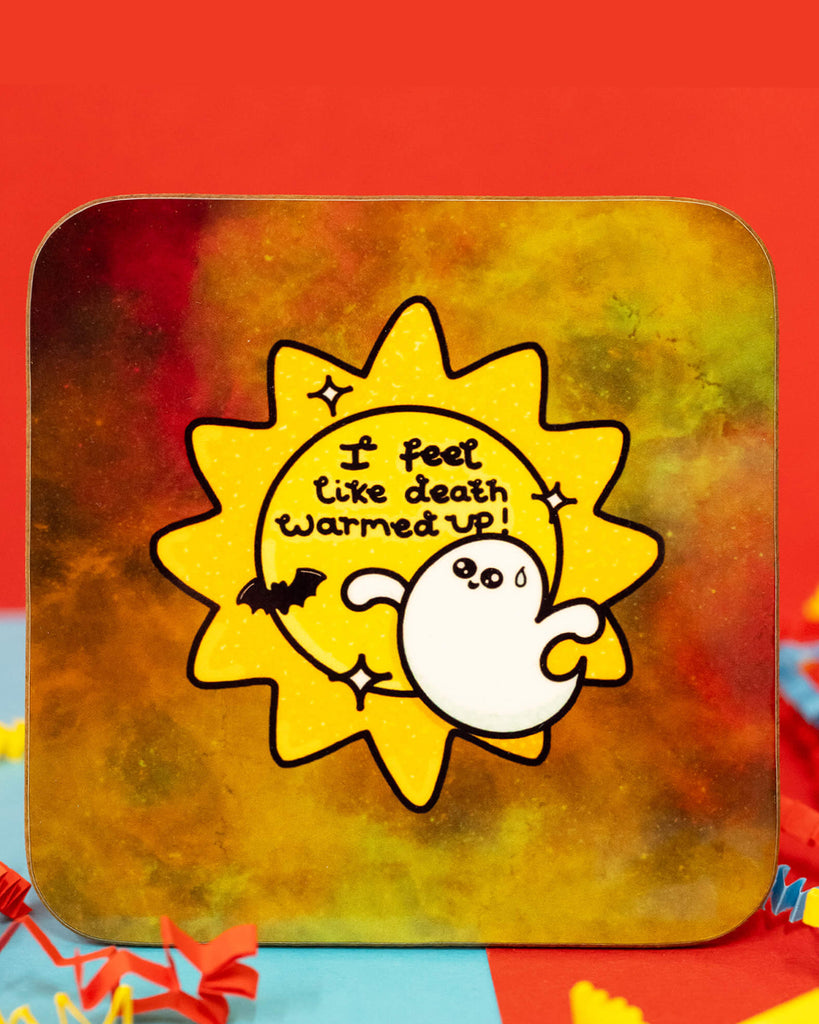 Close-up of the Death Warmed Up Coaster by Innabox. The square coaster has rounded corners and a mottled background in shades of yellow, orange, and green. The design features a worried-looking white ghost with a sweat drop, standing in front of a bright yellow sunburst with the phrase "I feel like death warmed up!" in playful black text. A tiny bat and star details surround the design. Colourful crinkle paper surrounds the coaster. Available on Rebel Romance