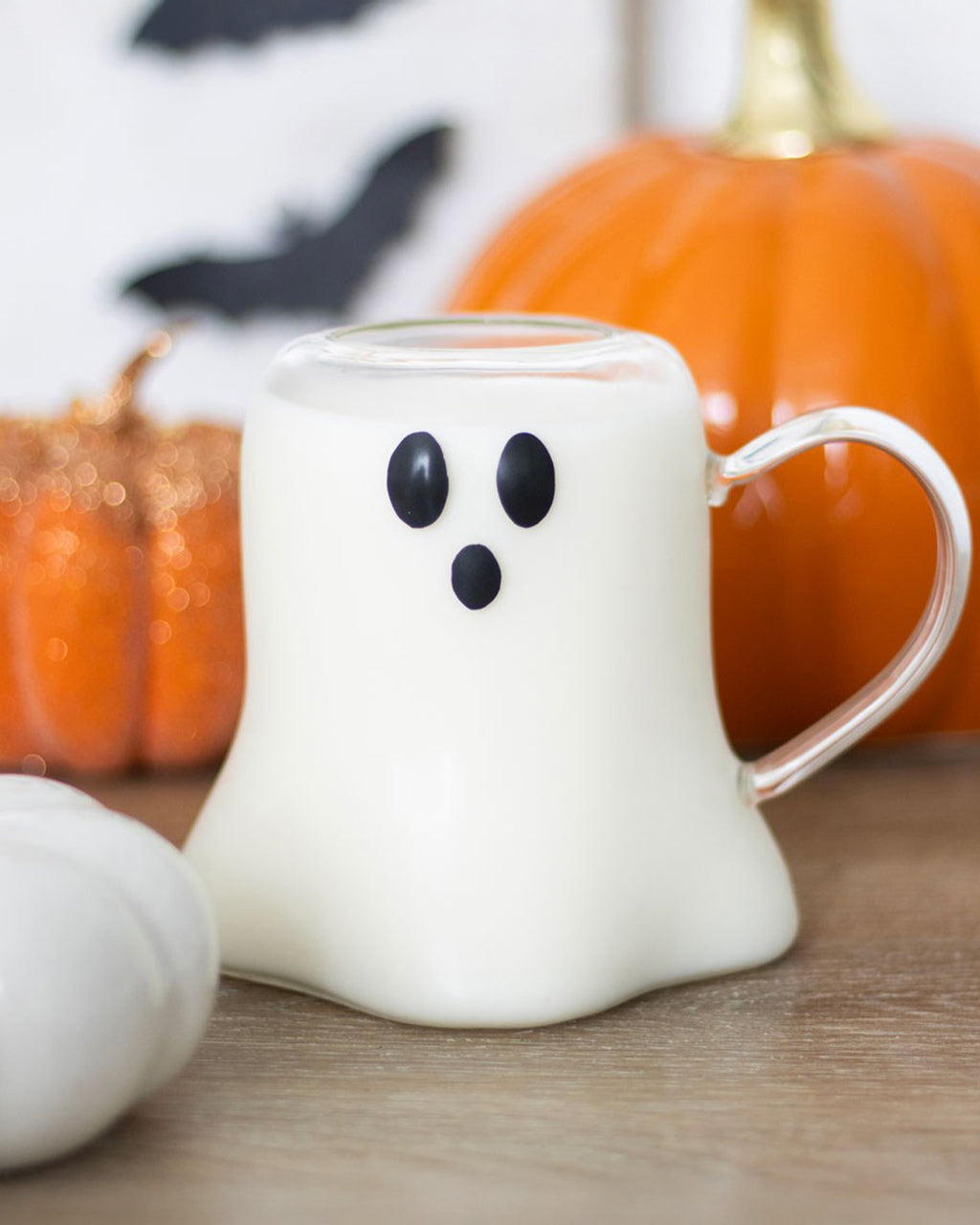 The Ghost Shaped Glass Mug from Rebel Romance filled with milk, creating an adorable ghost face effect. Set against a Halloween backdrop with pumpkins and bats, this mug is a must-have for spooky season drinks.