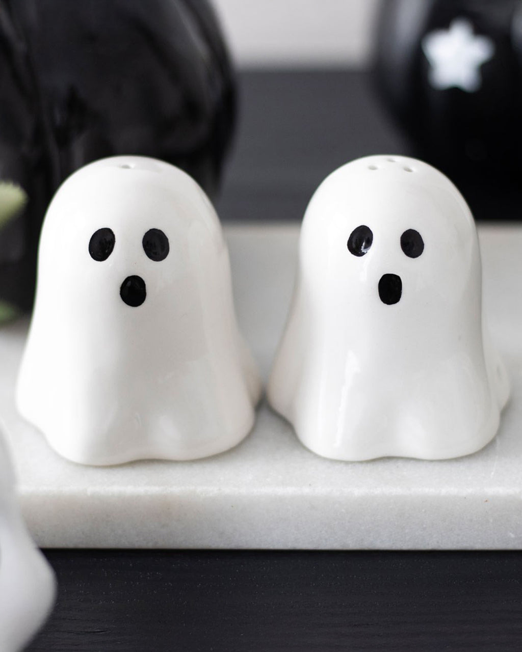 A spooky yet stylish kitchen setup featuring the Ghost Salt & Pepper Shaker Set from Rebel Romance. The shakers sit on a white tray, surrounded by halloween gothic decor, including black pumpkins and a dark wooden surface.
