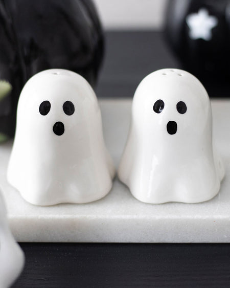 A spooky yet stylish kitchen setup featuring the Ghost Salt & Pepper Shaker Set from Rebel Romance. The shakers sit on a white tray, surrounded by halloween gothic decor, including black pumpkins and a dark wooden surface.