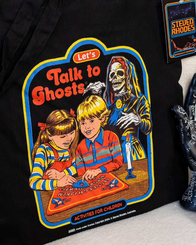 A black cotton tote bag featuring a retro-style Steven Rhodes design titled “Let’s Talk to Ghosts.” The illustration shows two children playing with a spirit board, with a skeleton ghost in the background. The vibrant design includes bold colours like red, yellow, and blue, paired with humorous text in a nostalgic comic-book style.