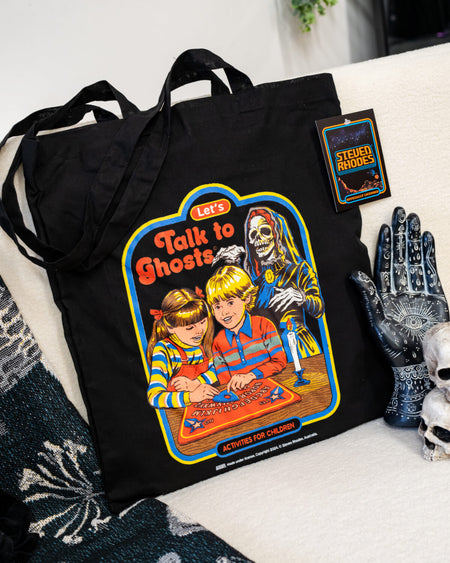 A black cotton tote bag featuring a retro-style Steven Rhodes design titled “Let’s Talk to Ghosts.” The illustration shows two children playing with a spirit board, with a skeleton ghost in the background. The vibrant design includes bold colours like red, yellow, and blue, paired with humorous text in a nostalgic comic-book style.