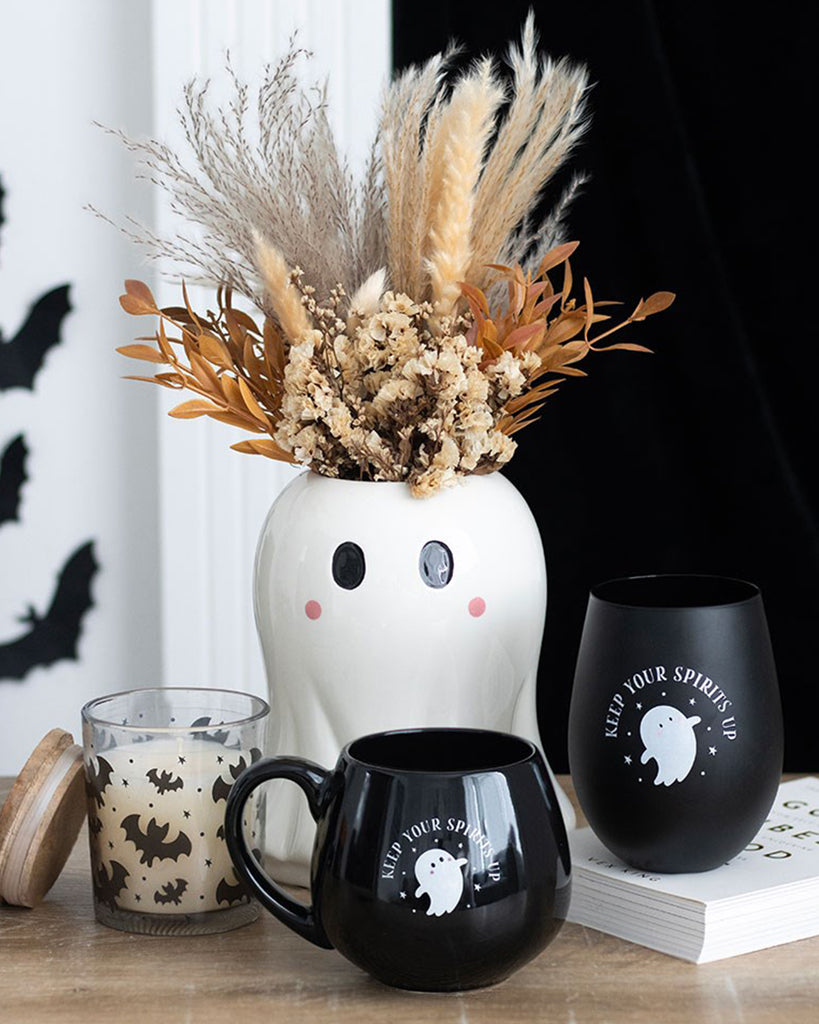 A ceramic ghost-shaped vase from Rebel Romance is styled with autumnal dried flowers, surrounded by spooky-themed black mugs and a bat-covered candle. Ideal for Halloween décor or cosy seasonal vibes.