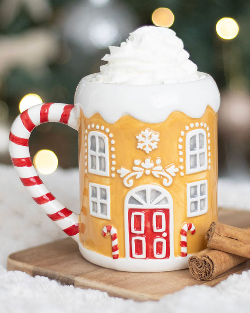 The Rebel Romance Candy Cane Gingerbread House Mug filled with whipped cream, sitting on a wooden board with cinnamon sticks in a cosy Christmas setting. A fun festive mug for winter nights and hot drinks.