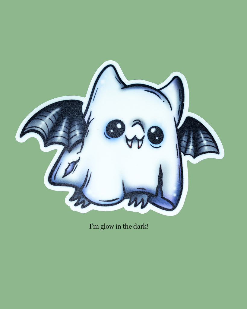 Glow in the dark ghost bat sticker featuring a cute cartoon bat dressed as a spooky ghost with torn edges and wide eyes. Black bat wings peek out from the ghost sheet design. Artwork by MilkTeef, perfect for Halloween lovers, spooky stationery collectors, and alternative style fans.