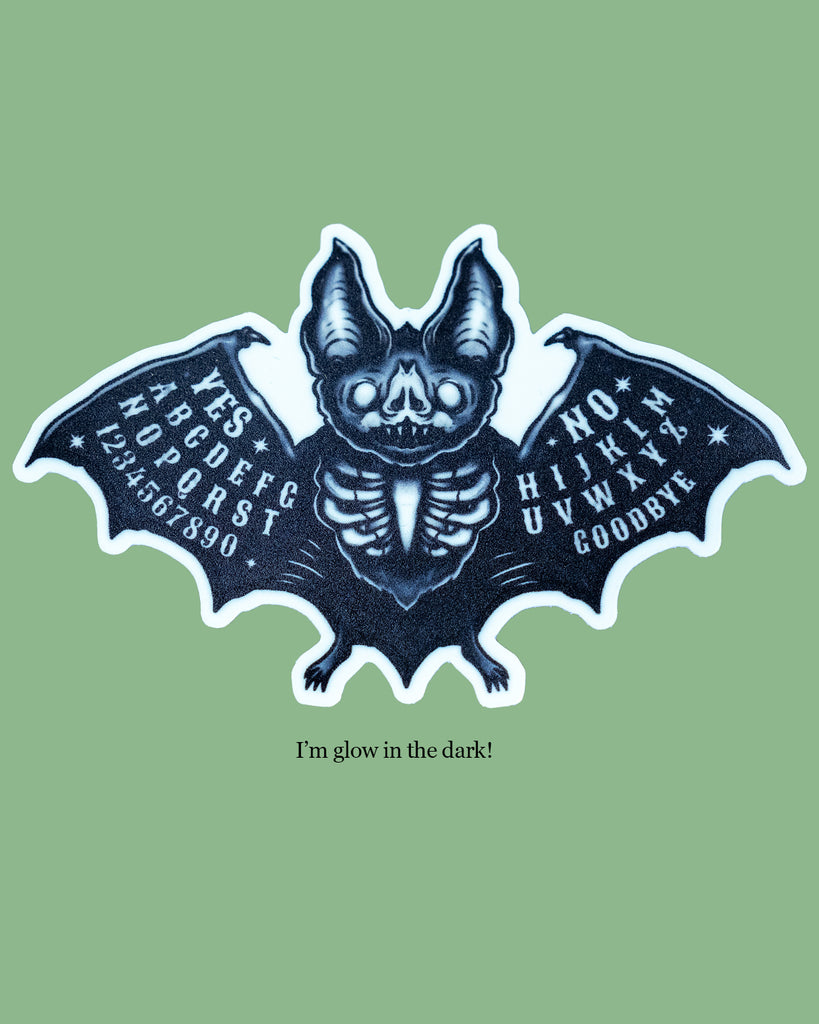 Glow in the dark Ouija bat sticker featuring spooky black and white bat with skeleton body and spirit board-inspired wings. Artwork by MilkTeef with YES, NO, letters, and numbers printed on each wing. Perfect gothic vinyl sticker for laptops, notebooks, and altars. Designed for Halloween and alternative style lovers.
