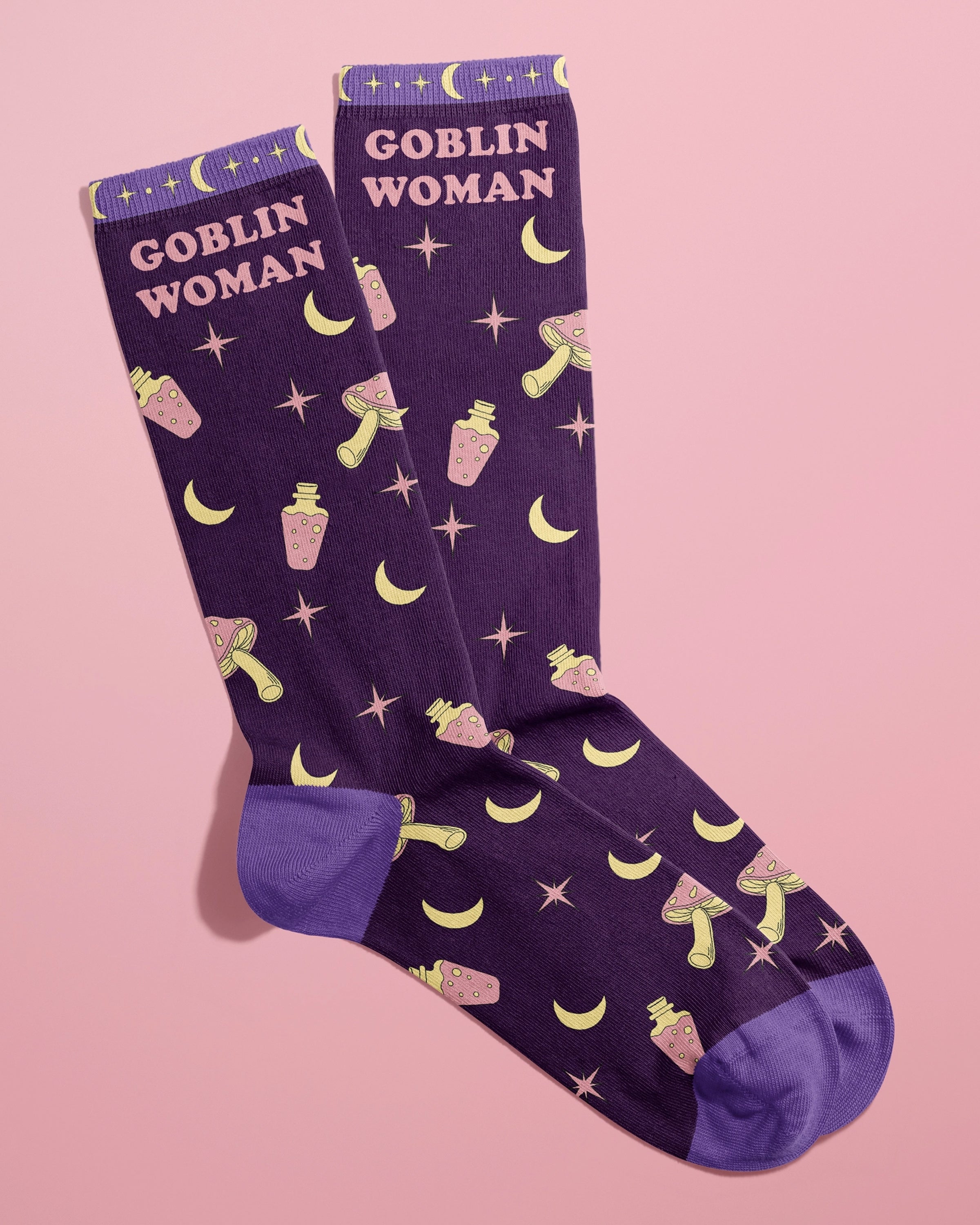 Pair of Rebel Romance Goblin Woman Socks by FUN CLUB styled together on a pastel pink background. The novelty socks feature a bold purple base with playful illustrations of yellow moons, pink potion bottles, stars, and mushrooms, and the words “Goblin Woman” in pastel pink. Great quirky gift idea for alternative fashion lovers.