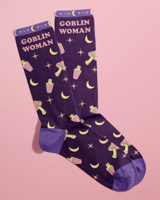 Pair of Rebel Romance Goblin Woman Socks by FUN CLUB styled together on a pastel pink background. The novelty socks feature a bold purple base with playful illustrations of yellow moons, pink potion bottles, stars, and mushrooms, and the words “Goblin Woman” in pastel pink. Great quirky gift idea for alternative fashion lovers.
