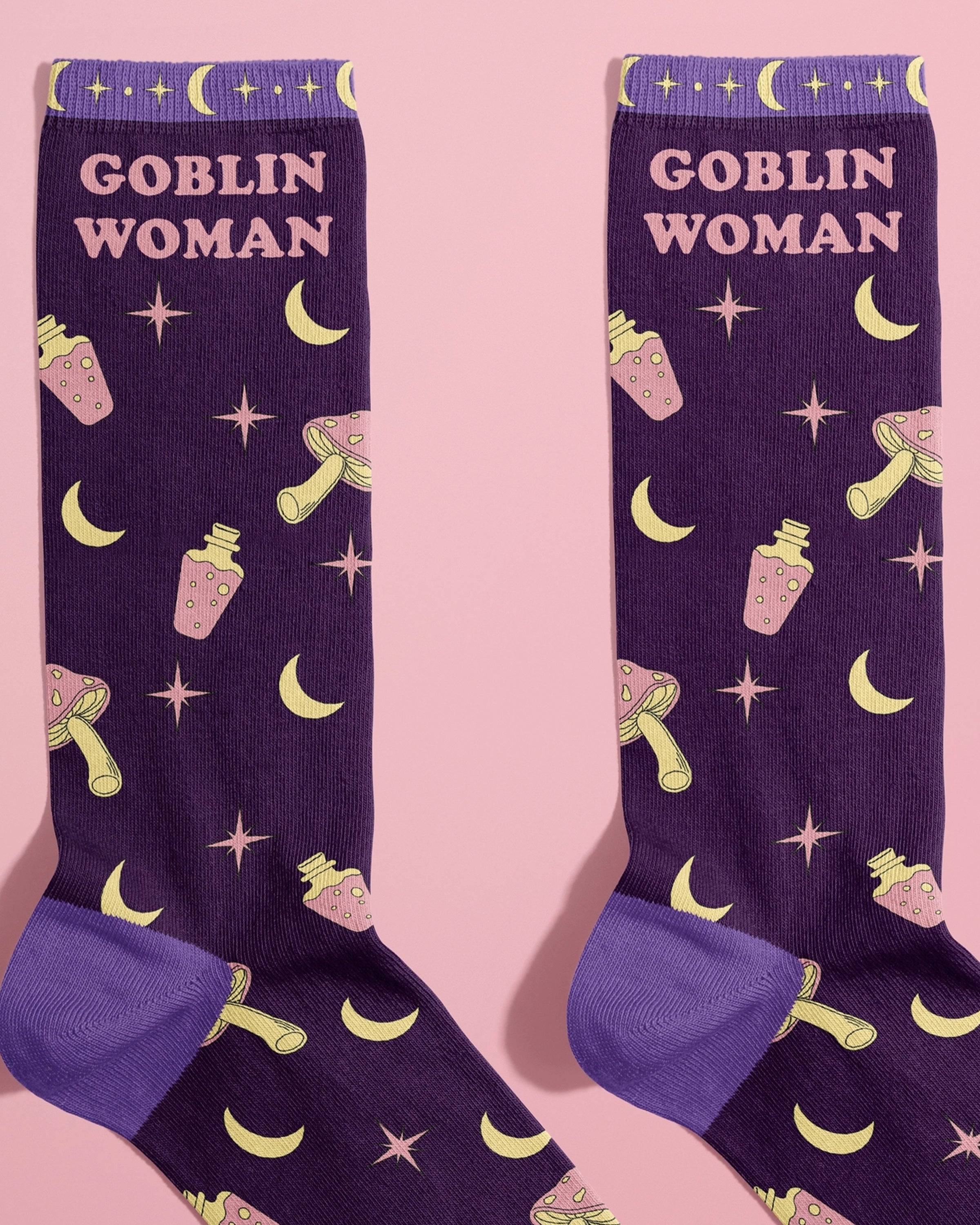 Flat lay of Rebel Romance’s Goblin Woman Socks by FUN CLUB, shown on a pastel pink background. The socks are deep purple with lavender heels and toes, featuring yellow crescent moons, pink potions, mushrooms, and star symbols, with “Goblin Woman” text at the top. A fun alternative fashion gift for millennials who love whimsical, witchy styles.