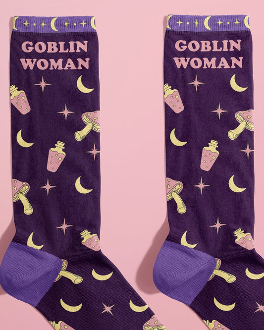 Flat lay of Rebel Romance’s Goblin Woman Socks by FUN CLUB, shown on a pastel pink background. The socks are deep purple with lavender heels and toes, featuring yellow crescent moons, pink potions, mushrooms, and star symbols, with “Goblin Woman” text at the top. A fun alternative fashion gift for millennials who love whimsical, witchy styles.