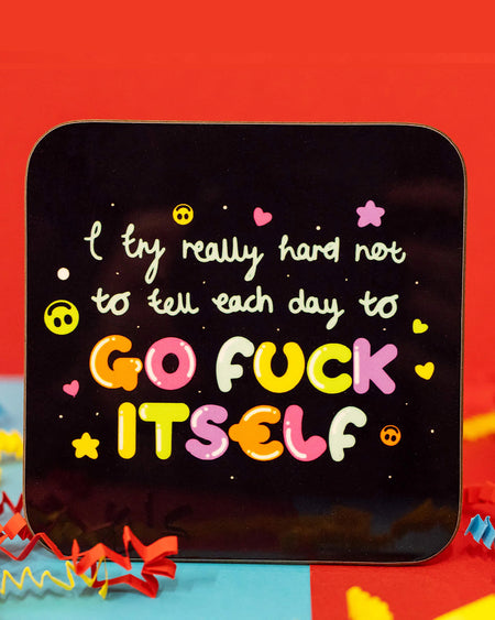 Close-up of the I Try Really Hard Coaster by Innabox. The square coaster has rounded corners and a glossy black background. The design features the phrase "I try really hard not to tell each day to GO F*** ITSELF" in playful, multicoloured bubble letters, with pink hearts, yellow smileys, stars, and dots scattered around. Colourful red, blue, and yellow crepe paper surrounds the coaster. Available on Rebel Romance