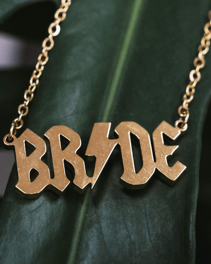 A flat lay of the gold-toned “BRIDE” necklace resting on a textured green leaf. The pendant’s shiny surface reflects light, emphasizing its bold design.