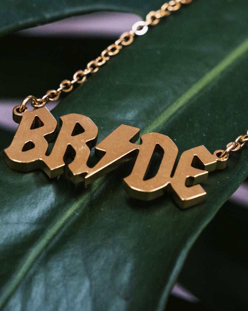 A flat lay of the gold-toned “BRIDE” necklace resting on a textured green leaf. The pendant’s shiny surface reflects light, emphasizing its bold design.