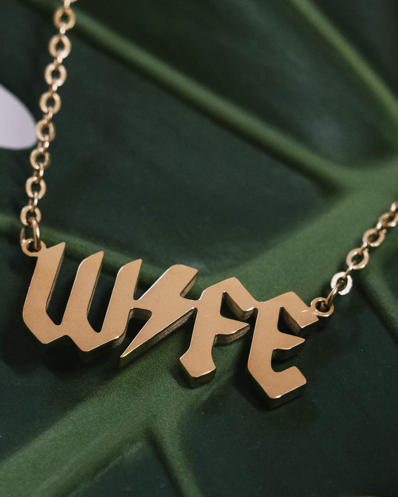 A gold-tone necklace with the word “WIFE” in bold, gothic-style lettering. The pendant rests on a lush green leaf, showcasing the necklace’s sleek and shiny finish.