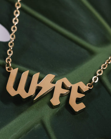 A gold-tone necklace with the word “WIFE” in bold, gothic-style lettering. The pendant rests on a lush green leaf, showcasing the necklace’s sleek and shiny finish.