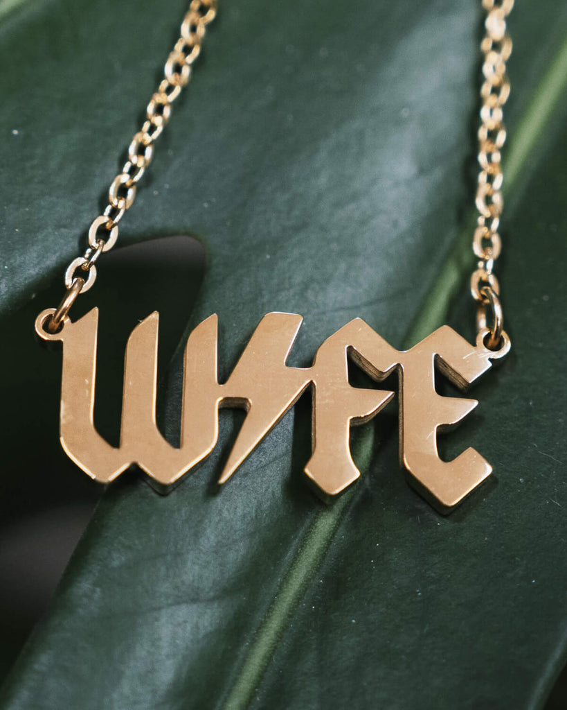 A gold-tone necklace with the word “WIFE” in bold, gothic-style lettering. The pendant rests on a lush green leaf, showcasing the necklace’s sleek and shiny finish.
