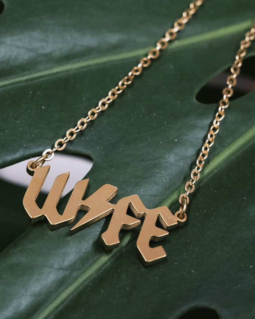 A gold-tone necklace with the word “WIFE” in bold, gothic-style lettering. The pendant rests on a lush green leaf, showcasing the necklace’s sleek and shiny finish.
