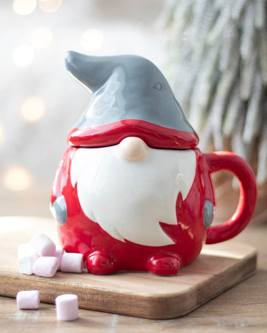 Festive ceramic gonk mug with its lid on, shaped like a gnome with red body, white beard and grey hat, standing next to pink marshmallows.