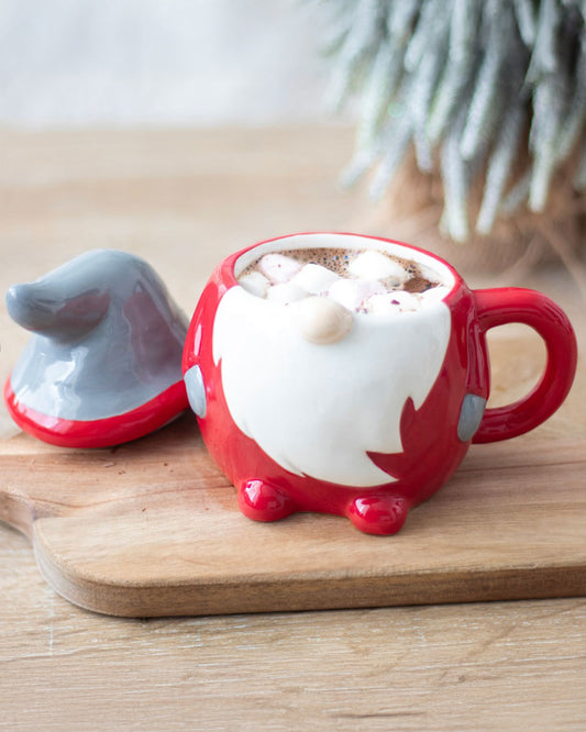 Red and grey ceramic Christmas gonk lidded mug filled with hot chocolate and marshmallows, styled on a wooden board with a frosted tree in the background.