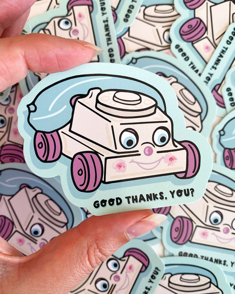 A close-up of a hand holding the "Creepy Phone" vinyl sticker stocked at Rebel Romance. The design features a retro toy-style telephone with a creepy-cute face, pink cheeks, purple wheels and a blue cord, with the caption “GOOD THANKS, YOU?” underneath. Designed by Yasmin Cousins and printed on durable vinyl.