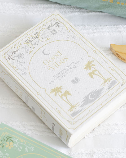 Good Vibes book-shaped storage box styled on a bed. Rebel Romance’s elegant faux book storage solution with celestial gold detailing blends seamlessly with home décor, making a unique gift or statement piece.