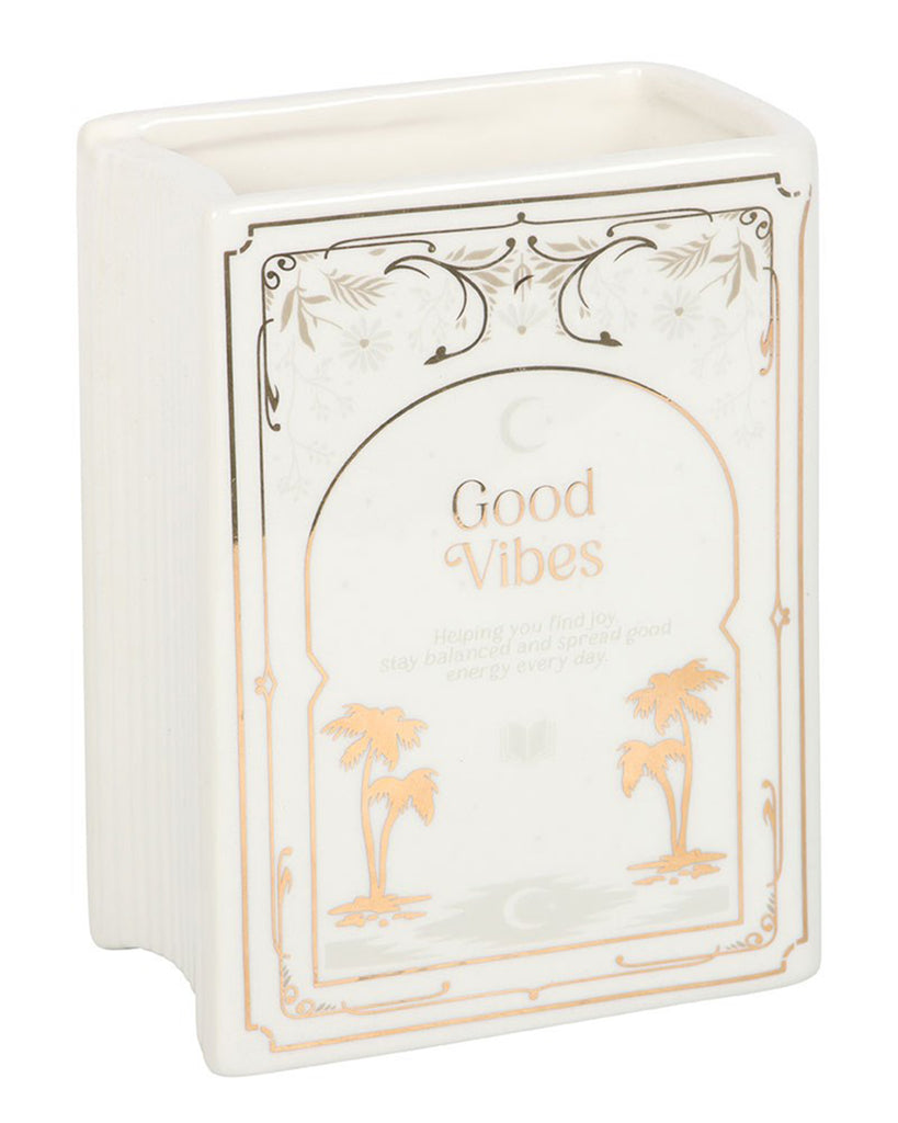 Angled view of Rebel Romance Good Vibes White Book Shaped Vase. This ceramic book-shaped vase in white with gold detailing adds charm to alternative home décor, making it perfect for book lovers and millennials seeking unique interior accessories.