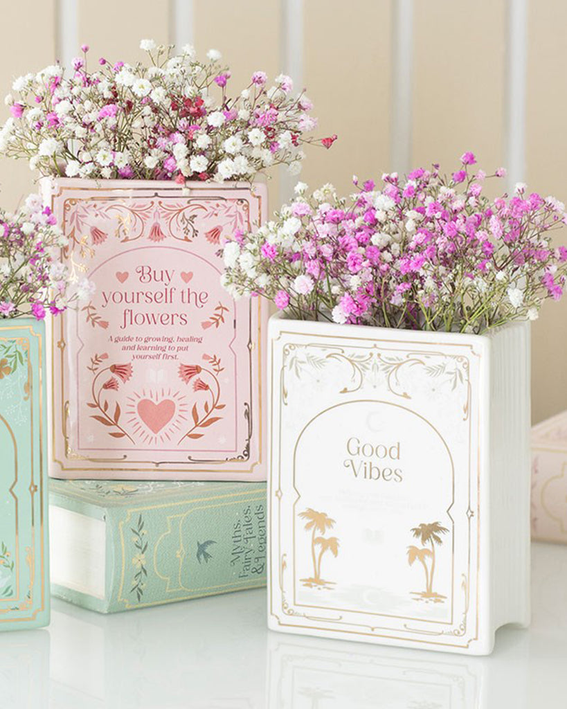 Rebel Romance Good Vibes White Book Shaped Vase styled with pink and white gypsophila flowers. This ceramic vase, designed to look like an upright book, makes a unique home accessory for book lovers, ideal for shelves, desks, or reading nooks. A whimsical gift for millennials who love alternative aesthetics.