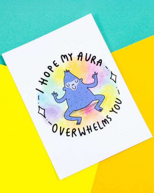 A white postcard of a smiling zen gorilla meditating illustration by Katie Abey with a squiggly rainbow background and black text reading "I hope my aura overwhelms you" written around it.