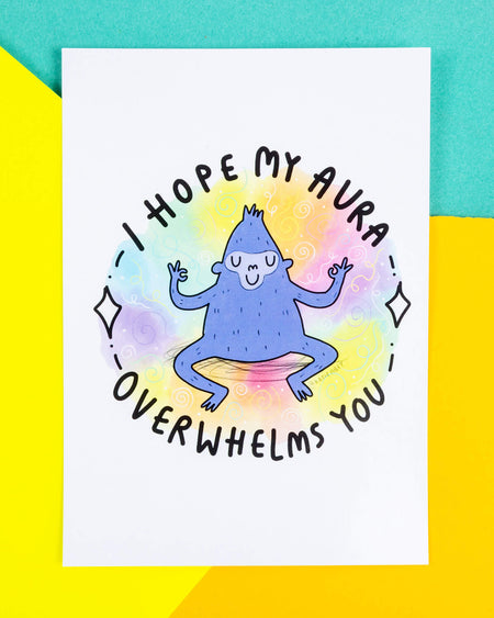 A white postcard of a smiling zen gorilla meditating illustration by Katie Abey with a squiggly rainbow background and black text reading "I hope my aura overwhelms you" written around it.